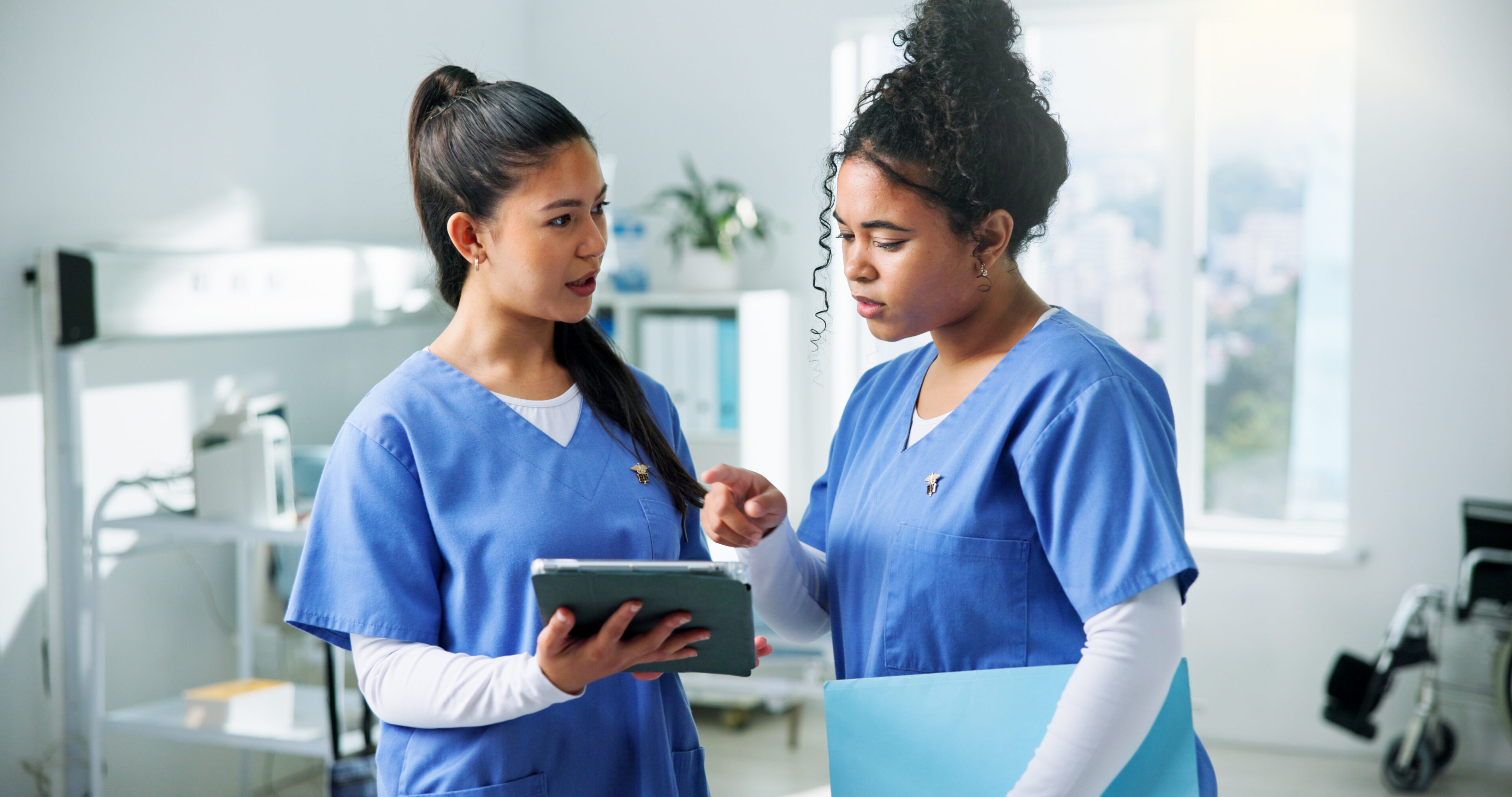 Two nurses in scrubs standing in a hospital room, looking at a tablet